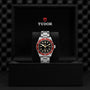 A TUDOR Black Bay wristwatch with a burgundy bezel, black dial, and silver metal band is shown in an open black TUDOR box set against a textured black background.