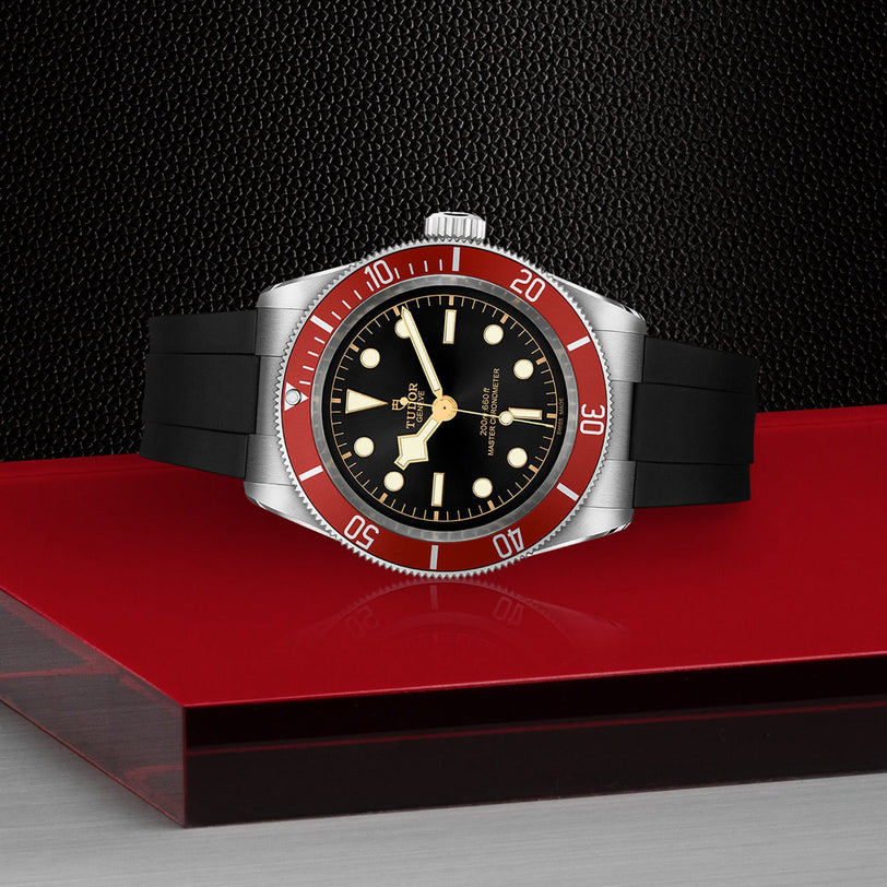 The TUDOR Black Bay wristwatch, featuring a burgundy bezel, black dial, and black rubber strap, is showcased on a red and black textured surface.
