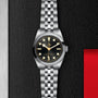 The TUDOR Black Bay 31, featuring a black dial and silver bracelet, exudes unisex style and is set against a textured black background with a red accent.