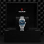The TUDOR Black Bay One 31, featuring a blue dial and silver bracelet, is displayed upright in a black presentation box with the TUDOR logo inside the lid, highlighting its unisex chic style and renowned Manufacture Calibres.