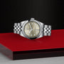 A TUDOR Black Bay One 31 in stainless steel with a champagne dial and bracelet is displayed on a glossy red and black surface, set against a textured black background.