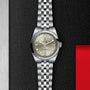 A TUDOR Black Bay One 31 wristwatch with a silver case, beige dial, and metal bracelet featuring the rapid adjustment system is displayed on a textured black surface beside a metallic and red background.