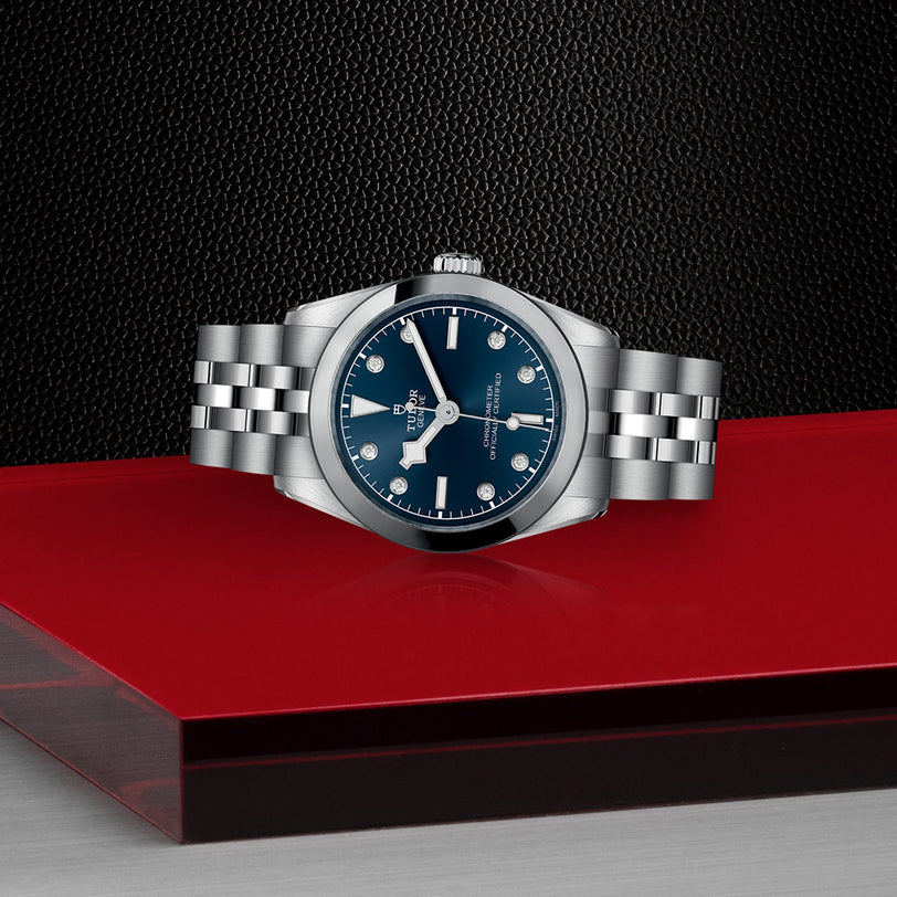 The TUDOR Black Bay One 31, a unisex stainless steel watch with a blue dial and silver bracelet, is showcased on a glossy red and black surface against a textured black background for a chic, sophisticated look.