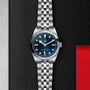 The TUDOR Black Bay One 31, a stainless steel wristwatch with a blue dial and metal bracelet, is showcased on a textured black surface with a red accent.