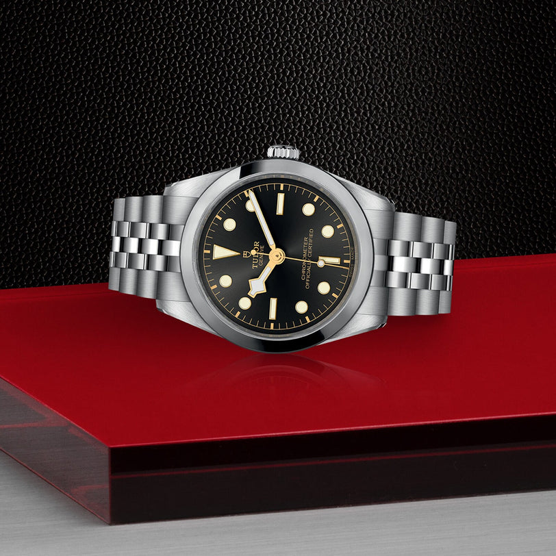 The TUDOR Black Bay One 36, a unisex stainless steel wristwatch with a black dial and jubilee bracelet, is showcased on a red and gray platform against a textured black background.