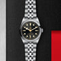 The TUDOR Black Bay One 36 unisex watch features a stainless steel case, black dial, and jubilee bracelet, showcased against a textured black and red backdrop.