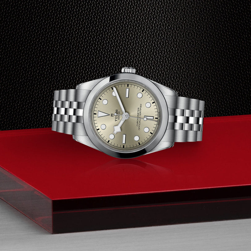 A TUDOR Black Bay One 36 unisex silver watch with a beige dial and metal bracelet is showcased on a red and black textured surface, reflecting the refined design of Black Bay models.