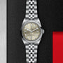 The TUDOR Black Bay One 36, a unisex silver watch with a cream dial and metal bracelet, is displayed on a textured black and red background, showcasing the timeless elegance of the brand’s advanced Manufacture Calibres.