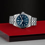 The TUDOR Black Bay One 36, a stainless steel unisex watch with a chic blue dial and metal bracelet inspired by Black Bay models, is displayed on a red and black geometric surface.
