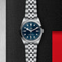 The TUDOR Black Bay One 36 wristwatch features a blue dial and silver metal bracelet, displayed against a textured black background.