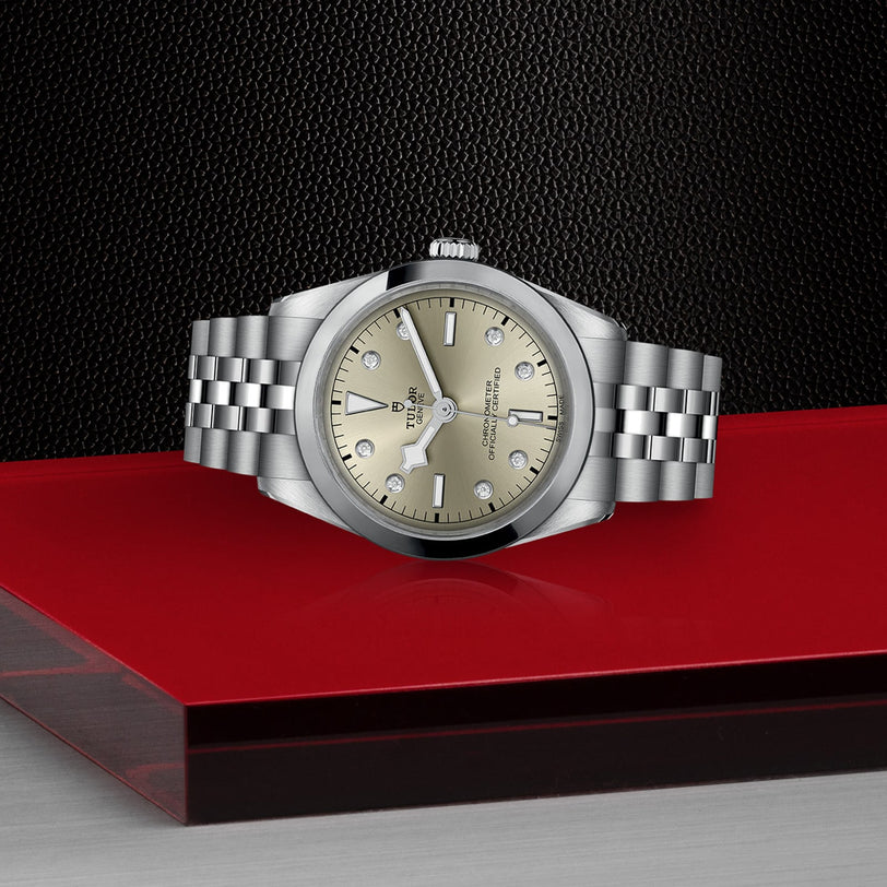 The TUDOR Black Bay One 36, with a stainless steel case, champagne dial, diamond hour markers, and 316L steel bracelet, is displayed on a red and black layered surface that highlights its elegant Black Bay design.