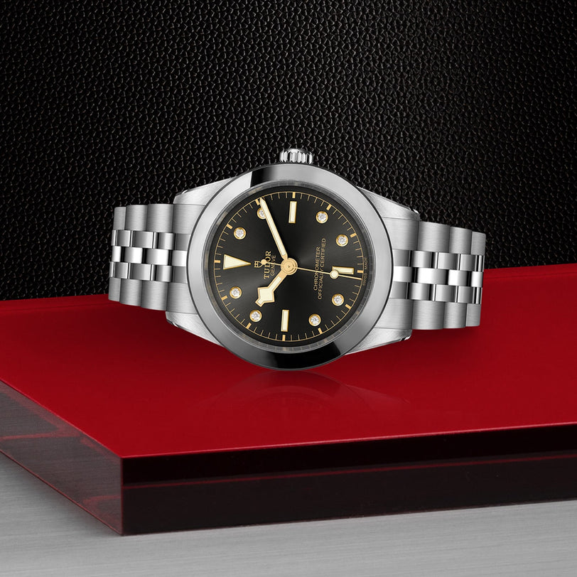 The TUDOR Black Bay One 39, a chic unisex stainless steel wristwatch with a black dial and gold hour markers, rests on a glossy red and black surface, reflecting its refined design against a textured black background.