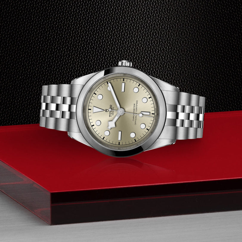 A TUDOR Black Bay One 41 wristwatch with a light gold dial and metal bracelet sits on red glass, radiating unisex style against a textured black backdrop.