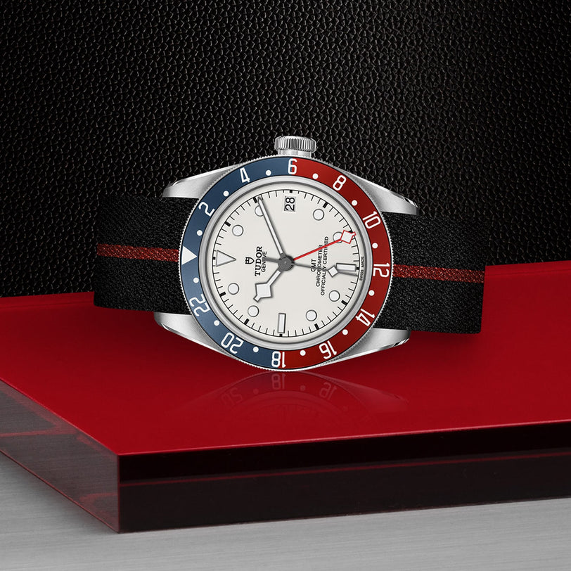 The TUDOR Black Bay GMT, featuring an opaline dial, blue and red bezel, and a black strap with a red stripe, is displayed on a striking red and black surface.