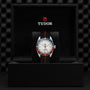 A TUDOR Black Bay GMT wristwatch with a red and blue bezel, black strap, and opaline dial stands upright in a black box bearing the TUDOR logo on the inner lid—ideal for stylishly tracking multiple time zones.