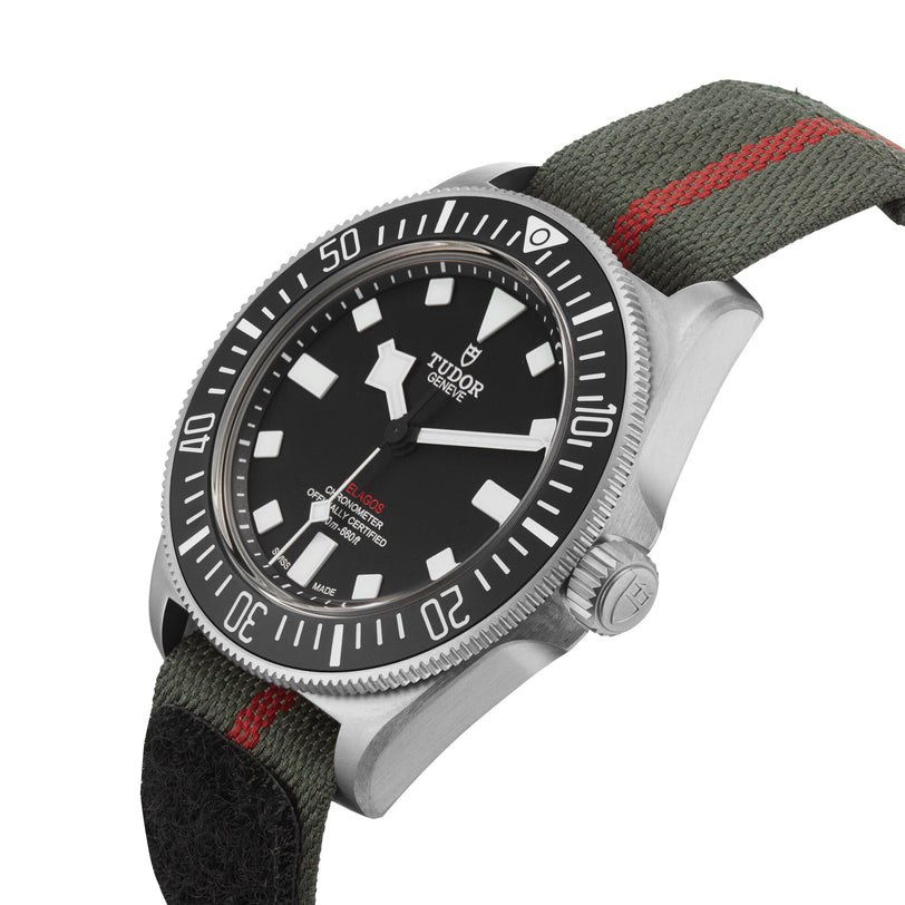 The TUDOR Pelagos FXD watch, featuring a black dial and bezel, white markers, and a green fabric strap with red stripe, is inspired by iconic TUDOR diving watches and is shown angled against a white background.