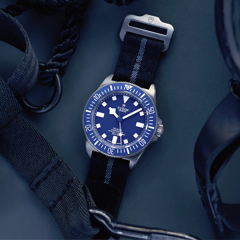 A TUDOR Pelagos FXD divers watch with a blue dial and NATO strap is displayed atop dark textured gear.