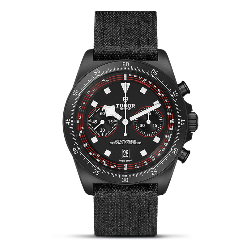 The TUDOR Pelagos FXD Chrono is a black wristwatch with a fabric strap, black bezel, cycling-specific tachymeter, white and red dial accents, two subdials, and a date display at 6 o’clock.
