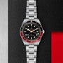 A TUDOR Black Bay 58 GMT dive watch with a black dial, red and black bezel, and metal bracelet sits on a textured black surface featuring a metallic edge and red section.