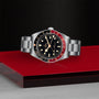 A TUDOR Black Bay 58 GMT watch with a black dial, red and black bezel, and metal bracelet rests on a red and silver surface against a textured black background.