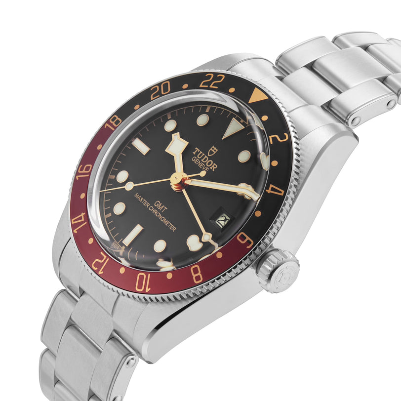 A TUDOR Black Bay 58 GMT wristwatch with a black dial, red and black bezel, and stainless steel bracelet is shown angled on a white background.
