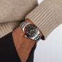 A person in a beige sweater and black pants wears a TUDOR Black Bay 58 GMT dive watch with a black and red bezel and black dial on their left wrist.