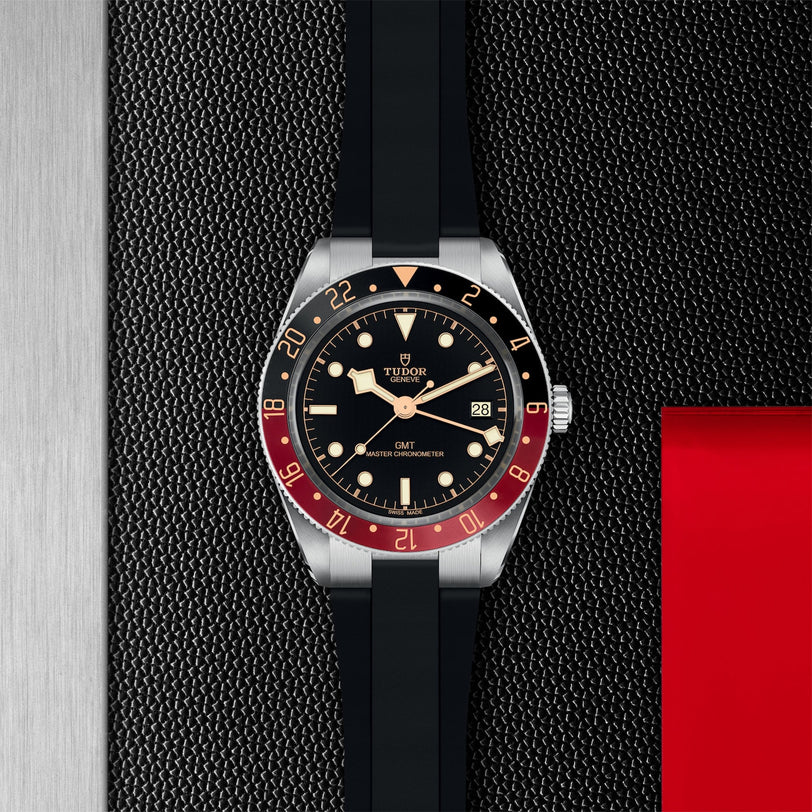The TUDOR Black Bay 58 GMT dive watch featuring a black dial, red and black bezel, rubber strap, and date display rests on a textured black surface next to metallic and red panels.