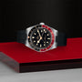 The TUDOR Black Bay 58 GMT, featuring a black dial, red and black bezel, and black rubber strap, is displayed on a red and metallic surface against a textured dark background.