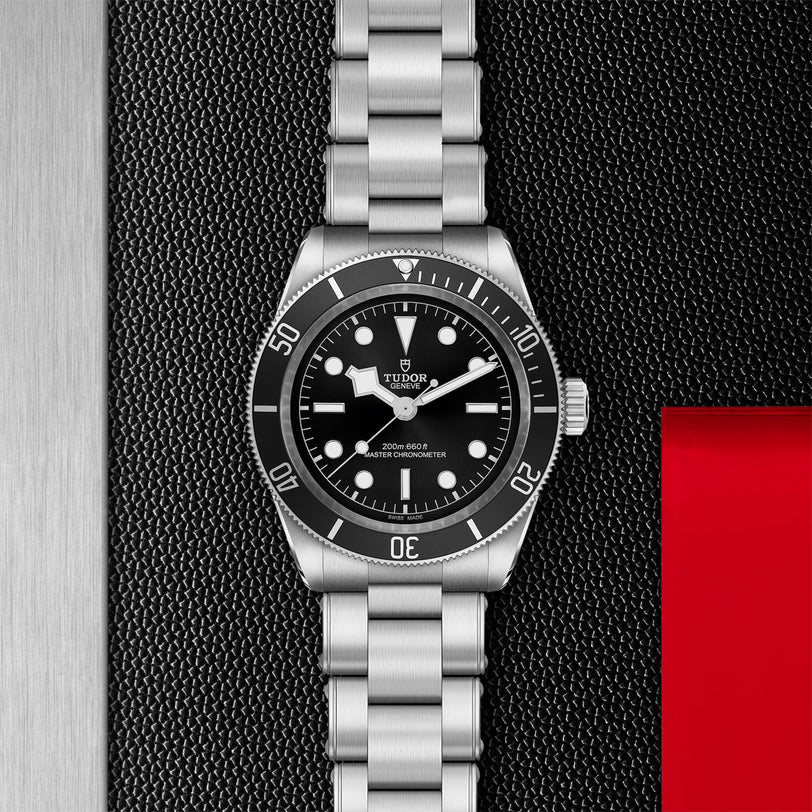 A TUDOR Black Bay watch featuring a stainless steel case, black dial and bezel, silver markers, and metal bracelet is showcased on a textured black background with a red accent—ideal for classic dive watch enthusiasts.