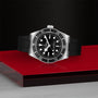 The TUDOR Black Bay is a METAS-certified stainless steel diving watch with a black dial, black bezel, black rubber strap, and white hour markers, shown on a red and silver surface against a dark textured background.