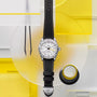 The TUDOR Black Bay Pro wristwatch with a black strap is displayed on a geometric yellow and white background with a magnifier nearby.