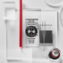 The TUDOR Black Bay Chrono wristwatch, featuring a sleek black dial and silver case, is showcased on a white and red geometric background with utensils.