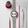 A TUDOR Black Bay Chrono wristwatch with a white dial is set on a gray surface between a loupe, a red tool, and tweezers.