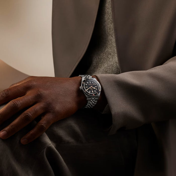 Black watch with dark dial and metal bracelet worn on wrist over brown sleeve.