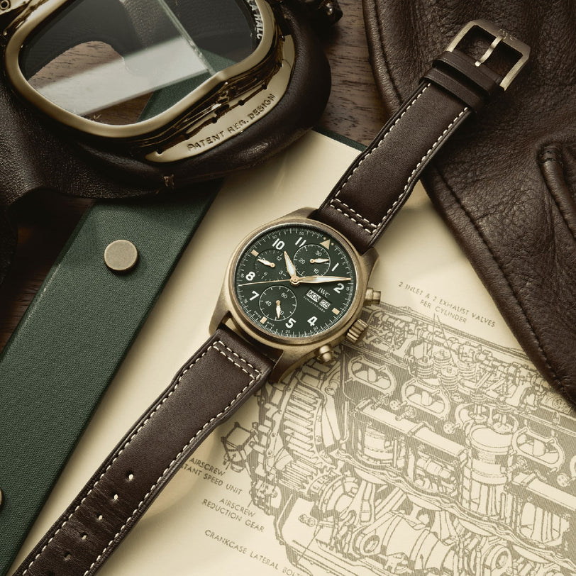 The IWC Schaffhausen Pilots Watch Chronograph Spitfire, with a green dial and brown leather strap, rests on a technical drawing. Its bronze case is shown beside aviator goggles and a brown leather jacket.