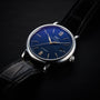The IWC Schaffhausen Portofino Automatic features a blue dial, silver hands and markers, a date window at 3 o’clock, and a black leather strap, exemplifying Swiss precision on a dark surface.
