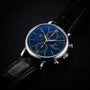 The IWC Schaffhausen Portofino Chronograph features a blue dial, gold hands and markers, three chronograph subdials, and a black leather strap, elegantly displayed on a dark surface.