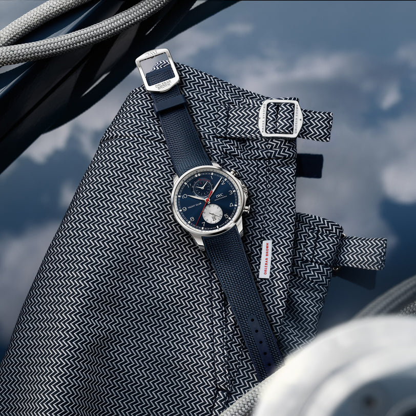 The IWC Schaffhausen Portugieser Yacht Club Chronograph Edition Orlebar Brown, with a blue strap, sits on patterned fabric amid metallic buckles and coiled ropes, reflected in water—a perfect pick for nautical sports watch enthusiasts.