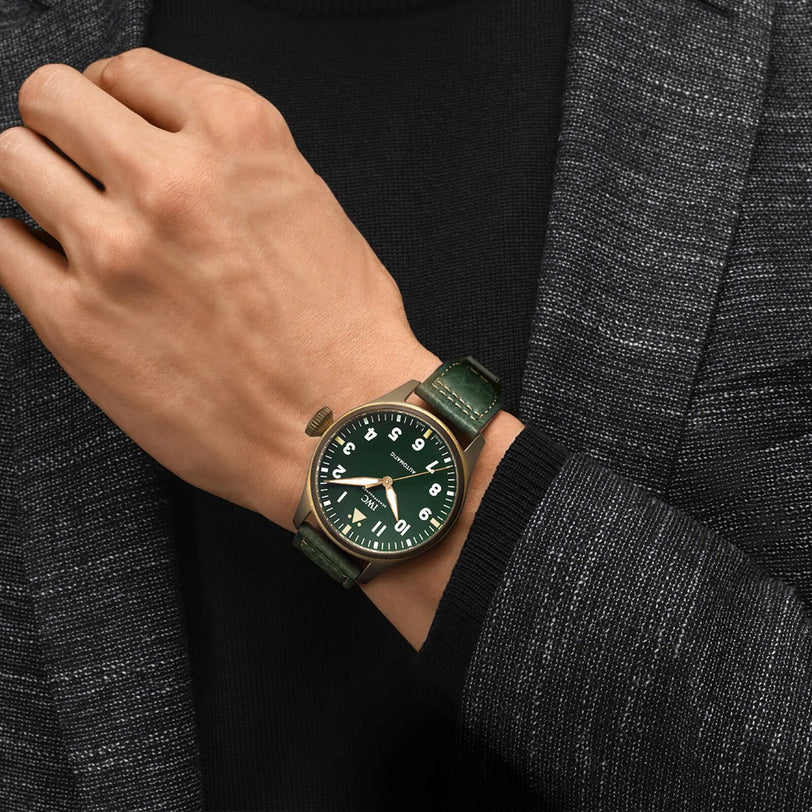 A person in a dark textured jacket wears an IWC Schaffhausen Big Pilots Watch 43 Spitfire with a green dial, bronze case, and EasX-CHANGE system on their left wrist.