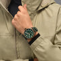 A person wearing a light beige jacket adjusts their collar with their left hand, showcasing the IWC Schaffhausen Big Pilots Watch 43 Spitfire—a wristwatch with a green dial, white numerals, and a bronze case.