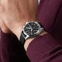 A person wearing an IWC Schaffhausen Pilots Watch Chronograph with automatic movement, a silver and black analog case, black dial, black leather strap, and a dark maroon long-sleeve shirt.