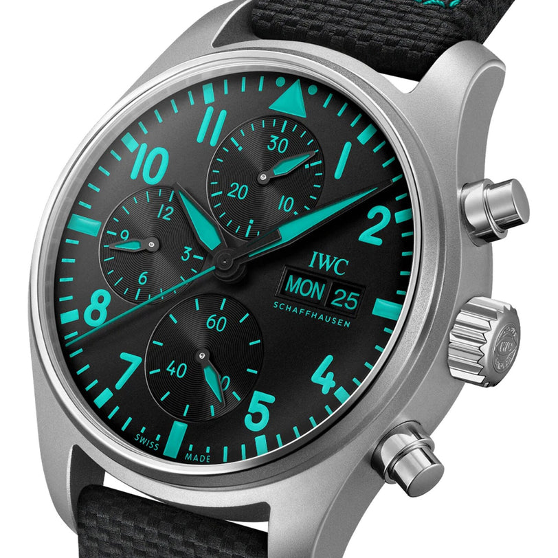 A close-up of the IWC Schaffhausen Pilots Watch Chronograph 41 Edition Mercedes-AMG PETRONAS Formula One™ Team with a black dial, turquoise luminous numerals and hands, three sub-dials, day/date display, fabric strap, and metal case.