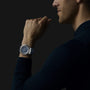 A person with light skin wears the Chopard Alpine Eagle XL Chrono—a silver Swiss watch with a dark face—while resting their chin on their hand against a dark background.