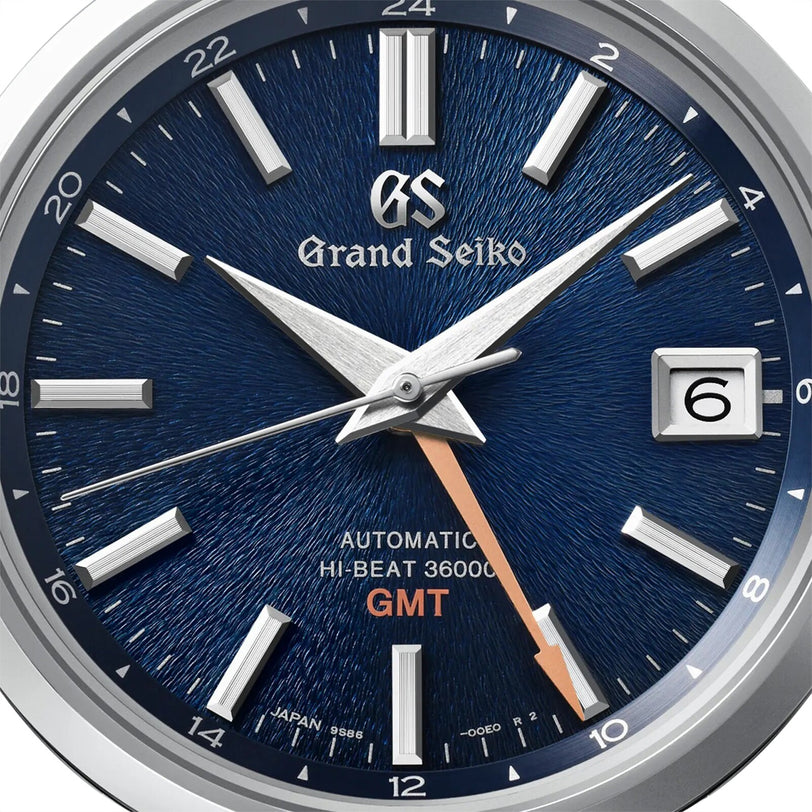 Close-up of a Grand Seiko Hi-Beat GMT Heritage watch face featuring a blue textured dial, silver hour markers, date window at 3 o’clock, and an orange GMT hand.