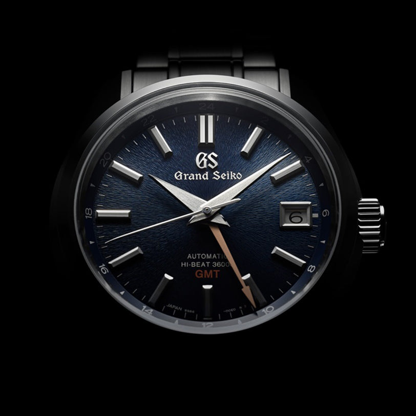 The Grand Seiko Hi-Beat GMT Heritage features Zaratsu polishing, a dark textured dial with silver markers and hands, a date window at 3 o’clock, and “GMT” in gold above 6 o’clock.