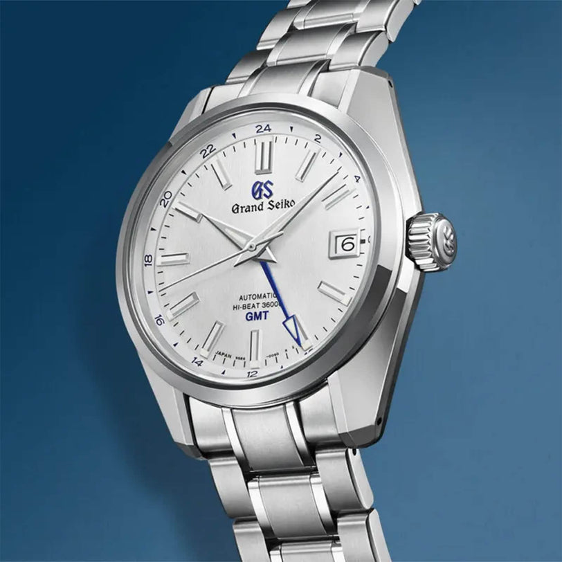 The Grand Seiko Hi-Beat GMT Heritage features stainless steel, a silver dial, date display, blue GMT hand, and 9S86 Hi-Beat 36 mechanical movement, shown against a blue background.