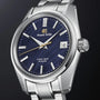 The Grand Seiko Hi-Beat Heritage wristwatch, handmade in Japan, features a blue patterned dial, silver hour markers, date window at 3 o’clock, and a stainless steel case and bracelet—precision in every detail.