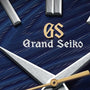 Close-up of the Grand Seiko Hi-Beat Heritage watch face, featuring a textured blue background, silver hands, gold second hand, and gold-and-white Grand Seiko logo—showcasing precision handcrafted in Japan.