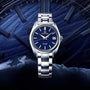 The Grand Seiko Hi-Beat Heritage, featuring a blue dial and stainless steel band, renowned for precision and handcrafted in Japan, is showcased against a stylized blue background with clock motifs and wave patterns.