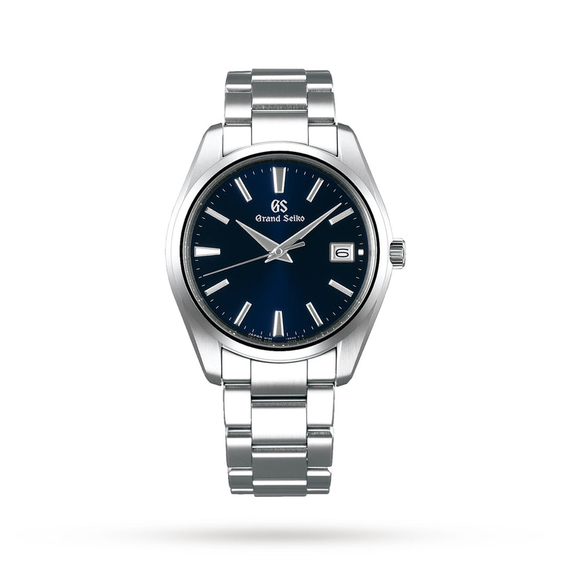 A Grand Seiko Quartz Heritage watch is shown on a white background, featuring a blue dial, silver hour markers, date at 3 o’clock, stainless steel case, and metal link bracelet.